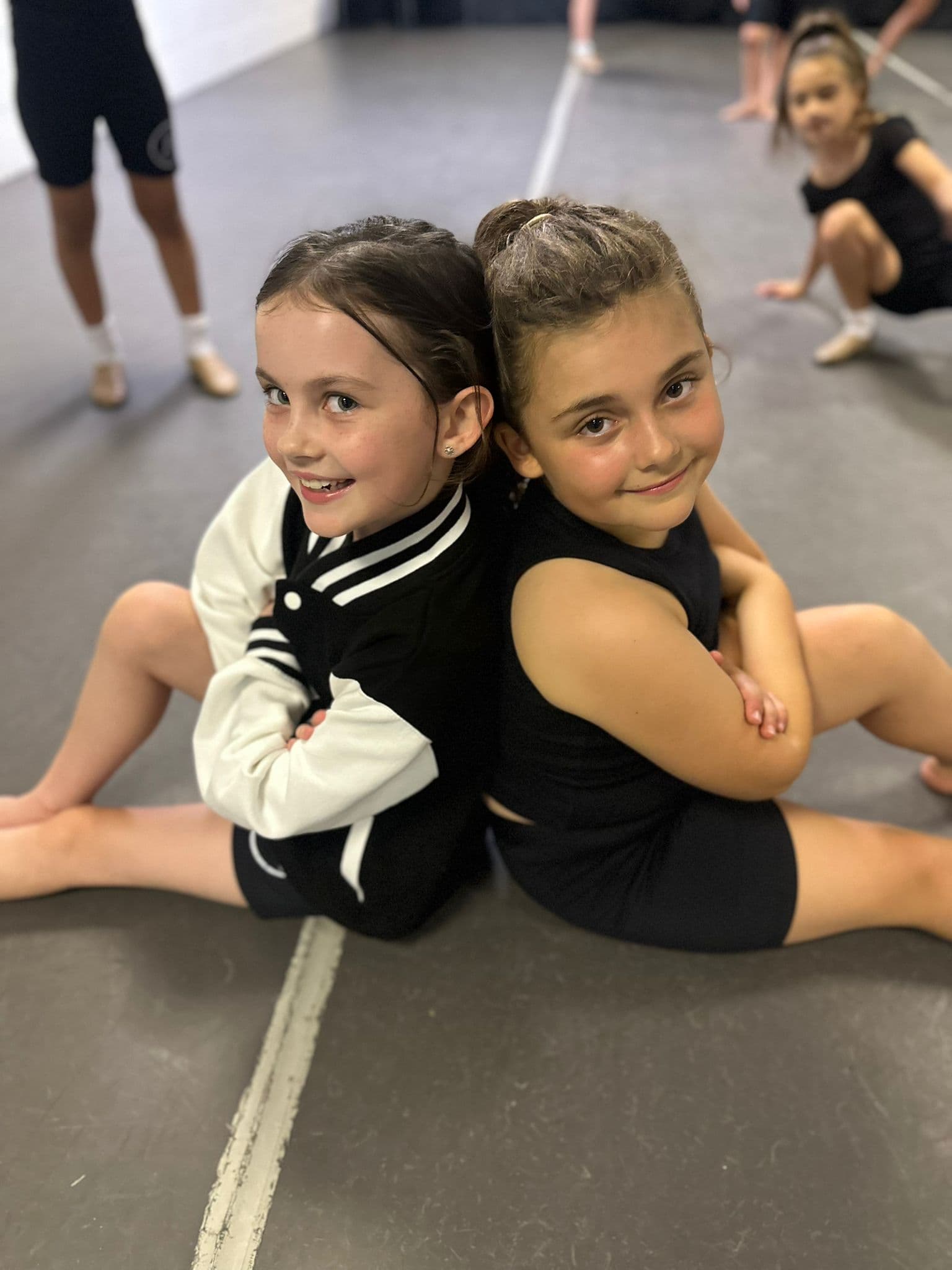 Two junior dancers sitting back to back, smiling in class